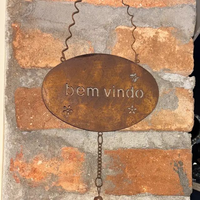 Placa de ferro com a frase “bem-vindo”, corrente e borboleta, pendurada numa parede de tijolinhos